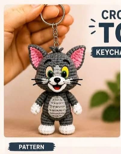 A crocheted gray amigurumi cat keychain with pink-lined ears and a white muzzle and paws, hanging from a keychain ring