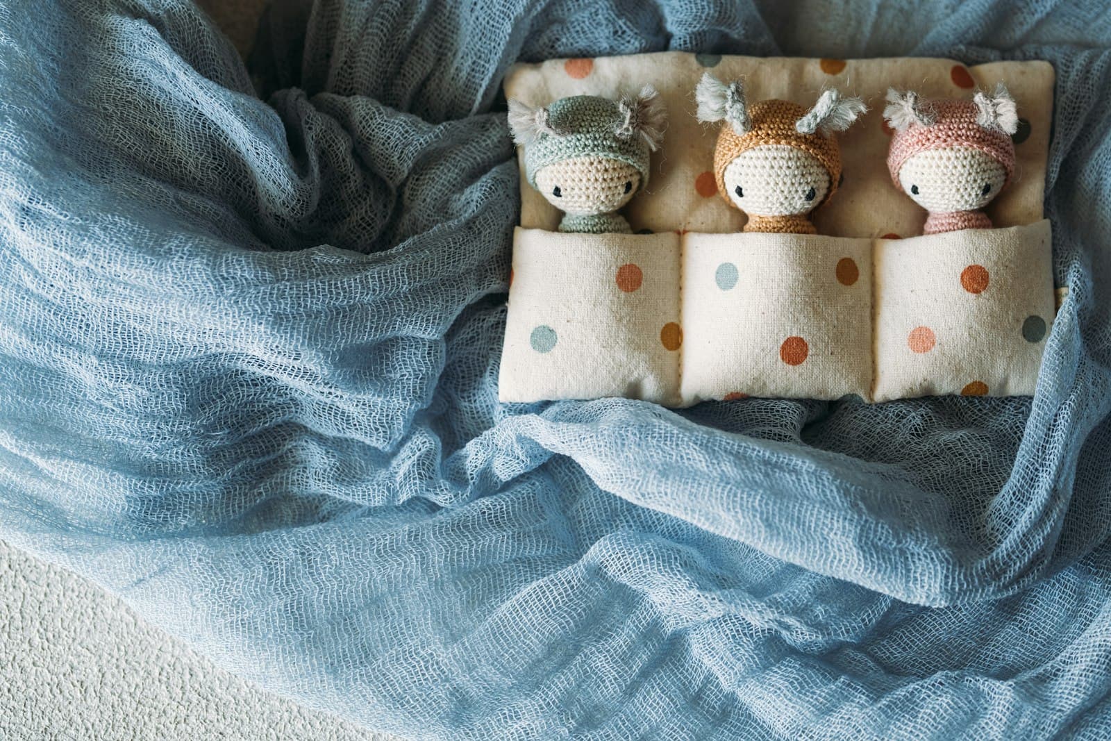 Three small amigurumi dolls with knitted hoods, propped in a fabric pocket against a blue blanket